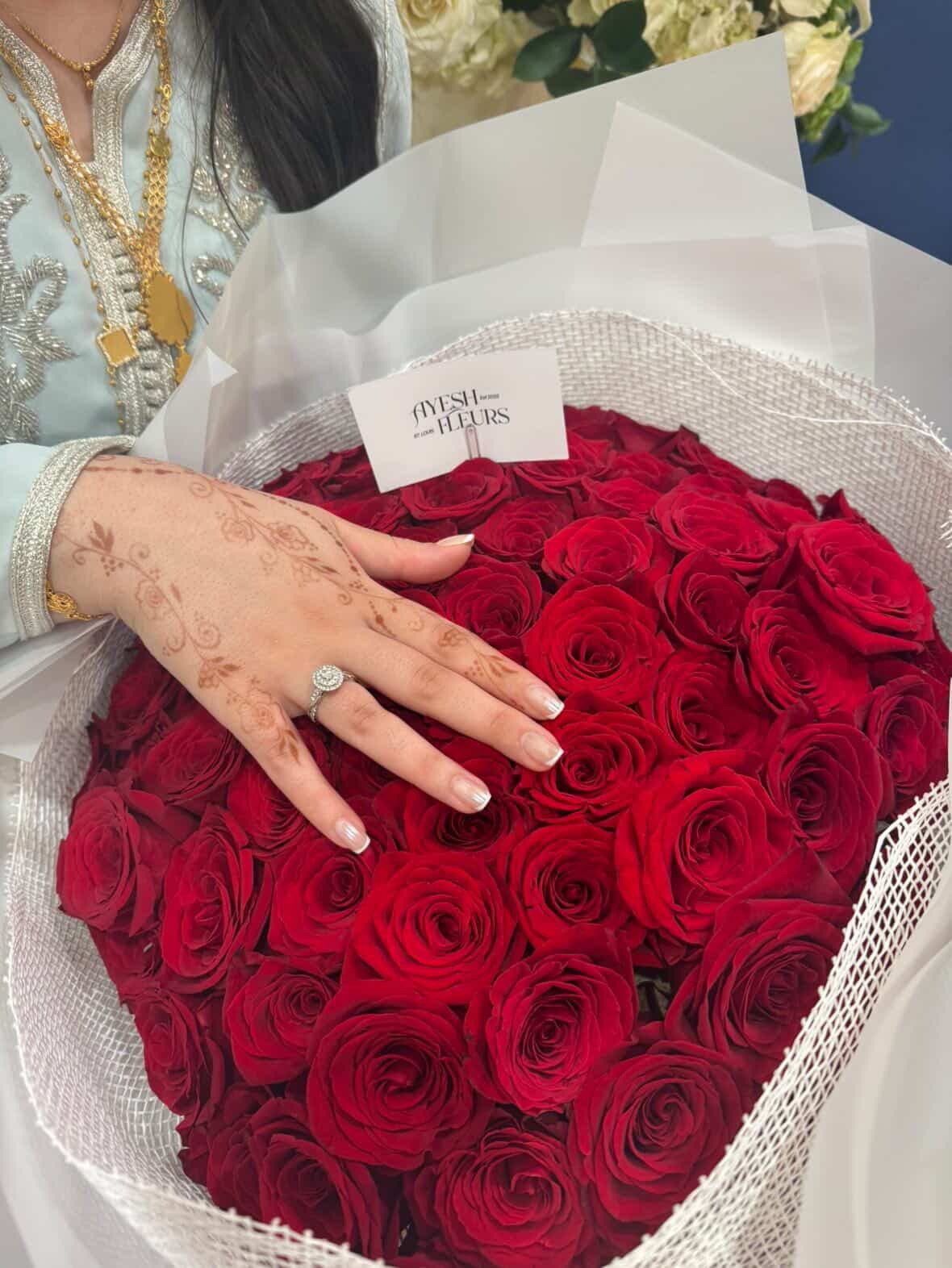 Round bouquet of red roses in white wrap with Ayesh Fleurs card, touched by a henna-decorated hand