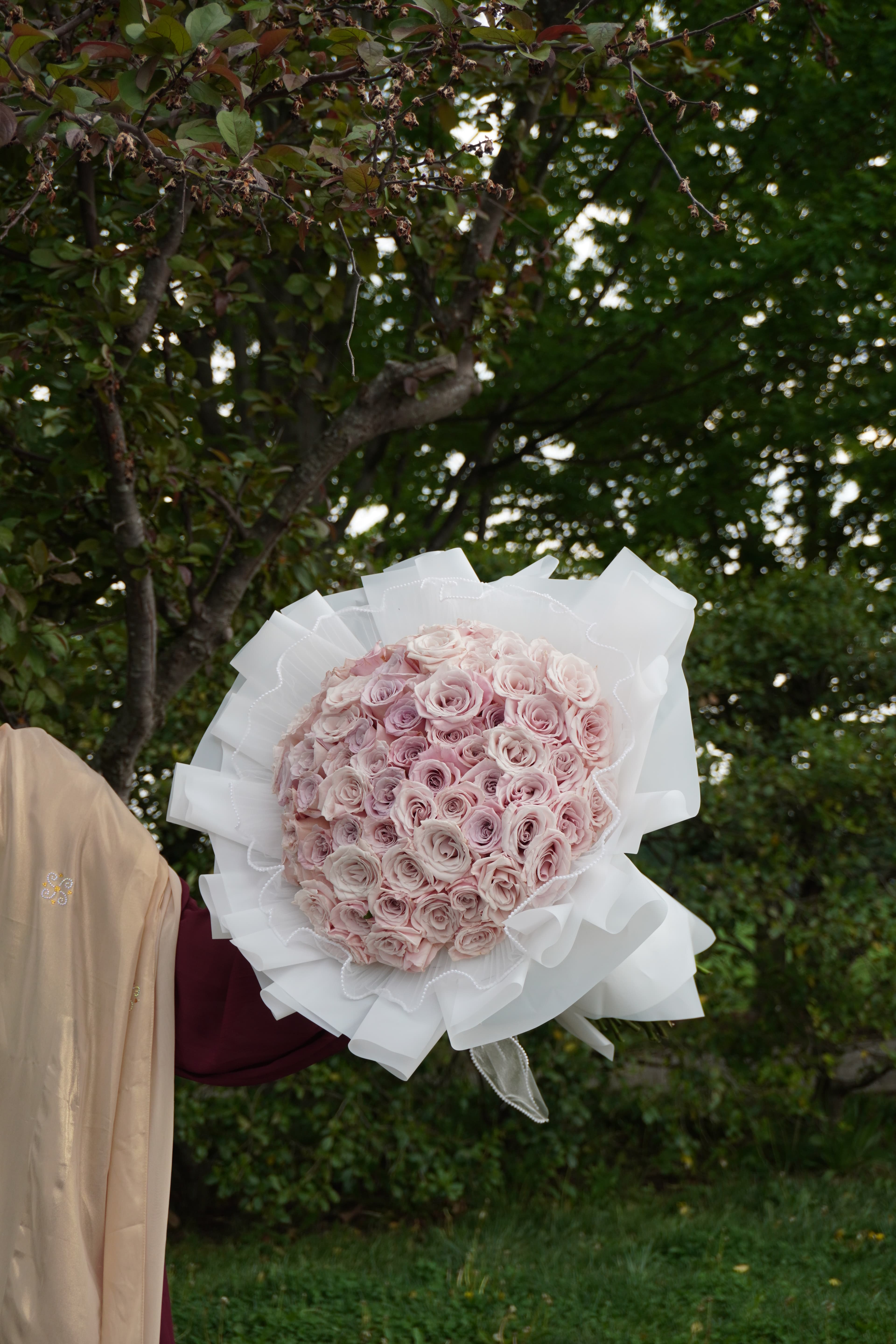 Round bouquet of blush pink roses in ruffled white wrap, held outdoors among green trees