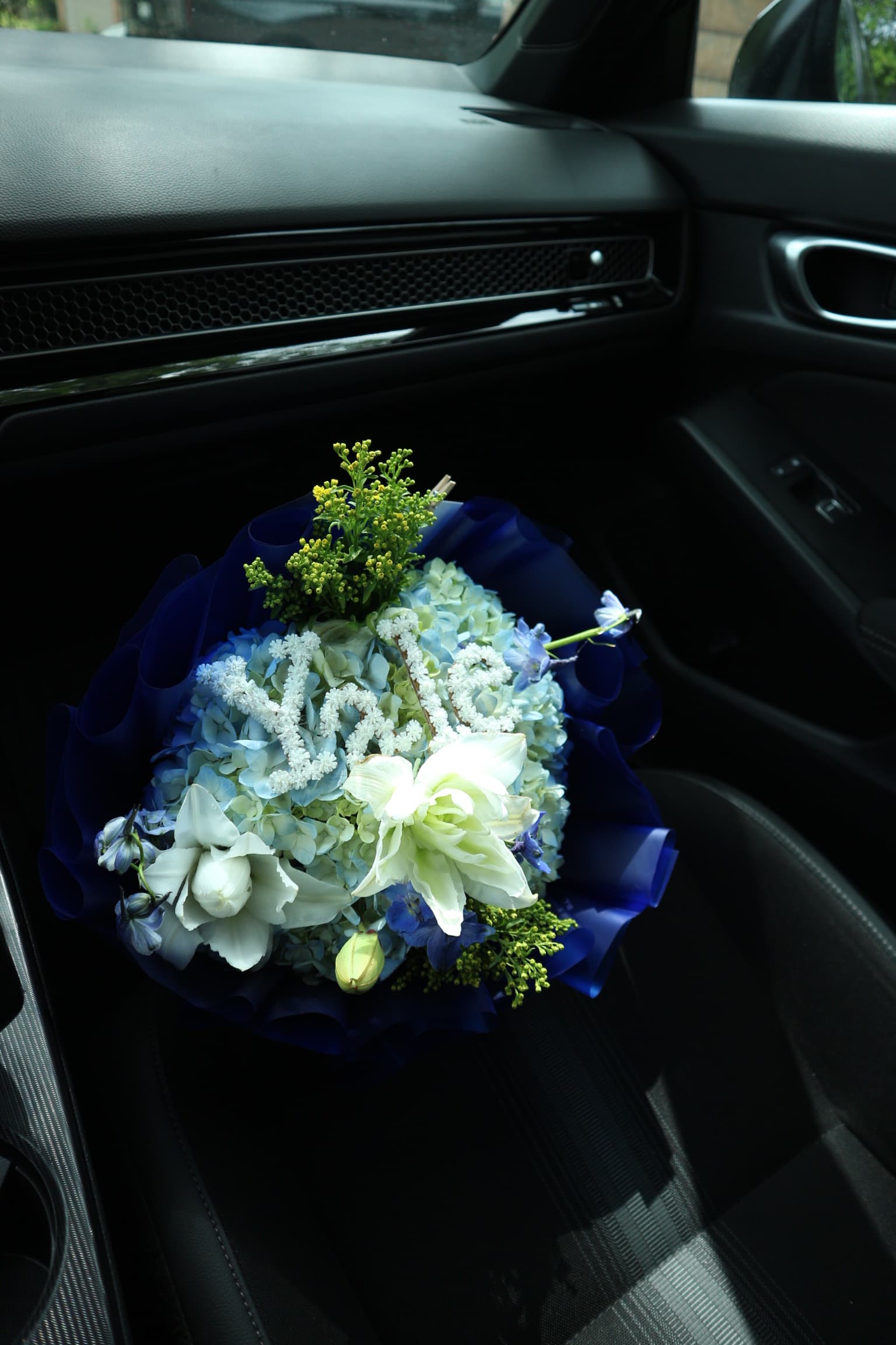 Blue hydrangea bouquet with white lilies and yellow solidago in royal blue wrap, resting on a car seat