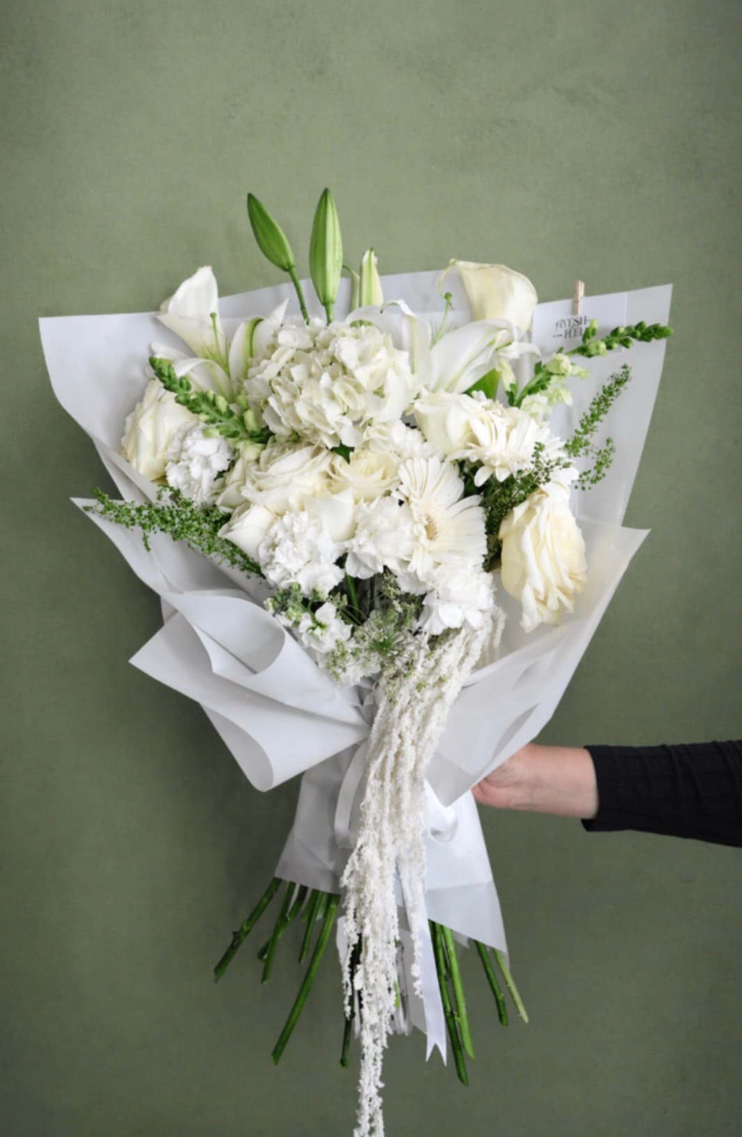 White bouquet of lilies, roses, gerbera and snapdragons with trailing amaranthus against a green wall