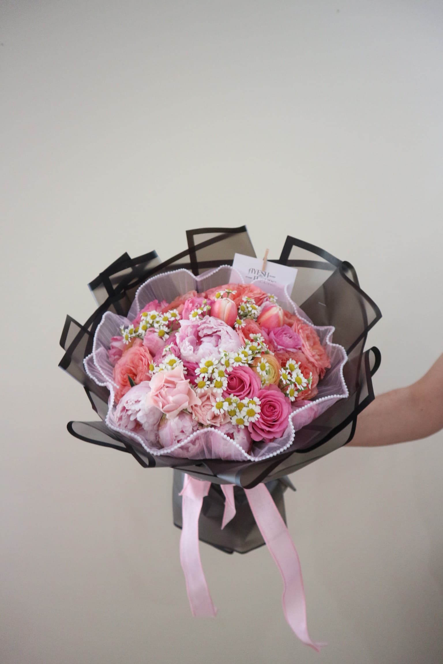 Pink peonies, roses, ranunculus and chamomile daisies in black tulle wrap with pink ribbon, held up