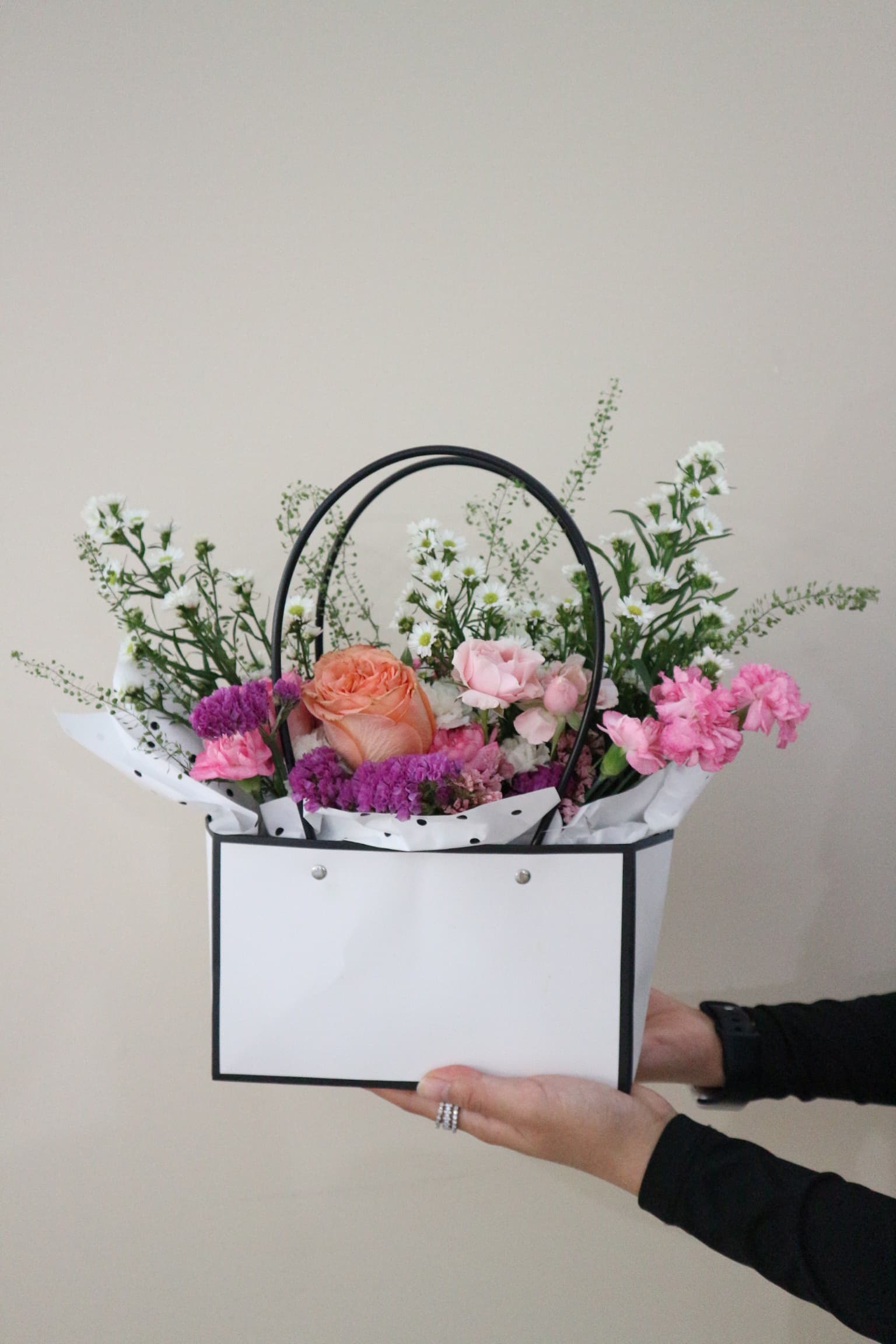 White floral bag with peach rose, pink carnations, purple statice and daisy sprays, held by a hand