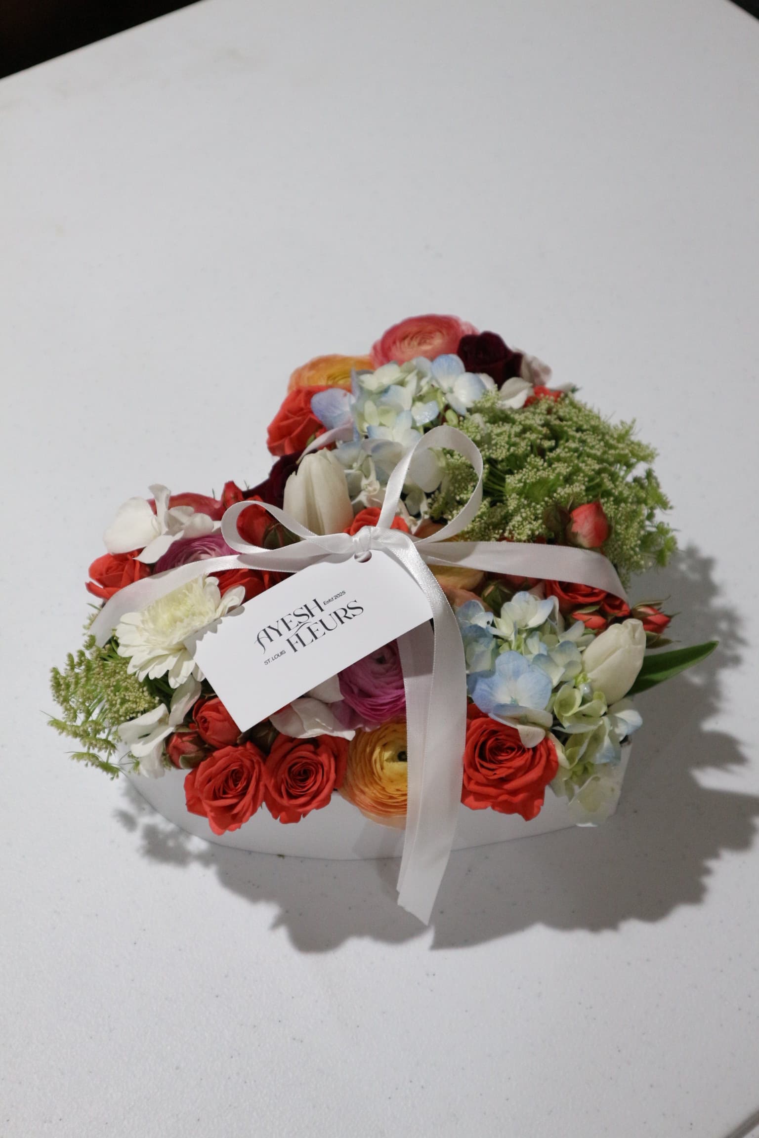 Heart box of red spray roses, ranunculus, white tulips and blue hydrangea tied with Ayesh Fleurs ribbon tag