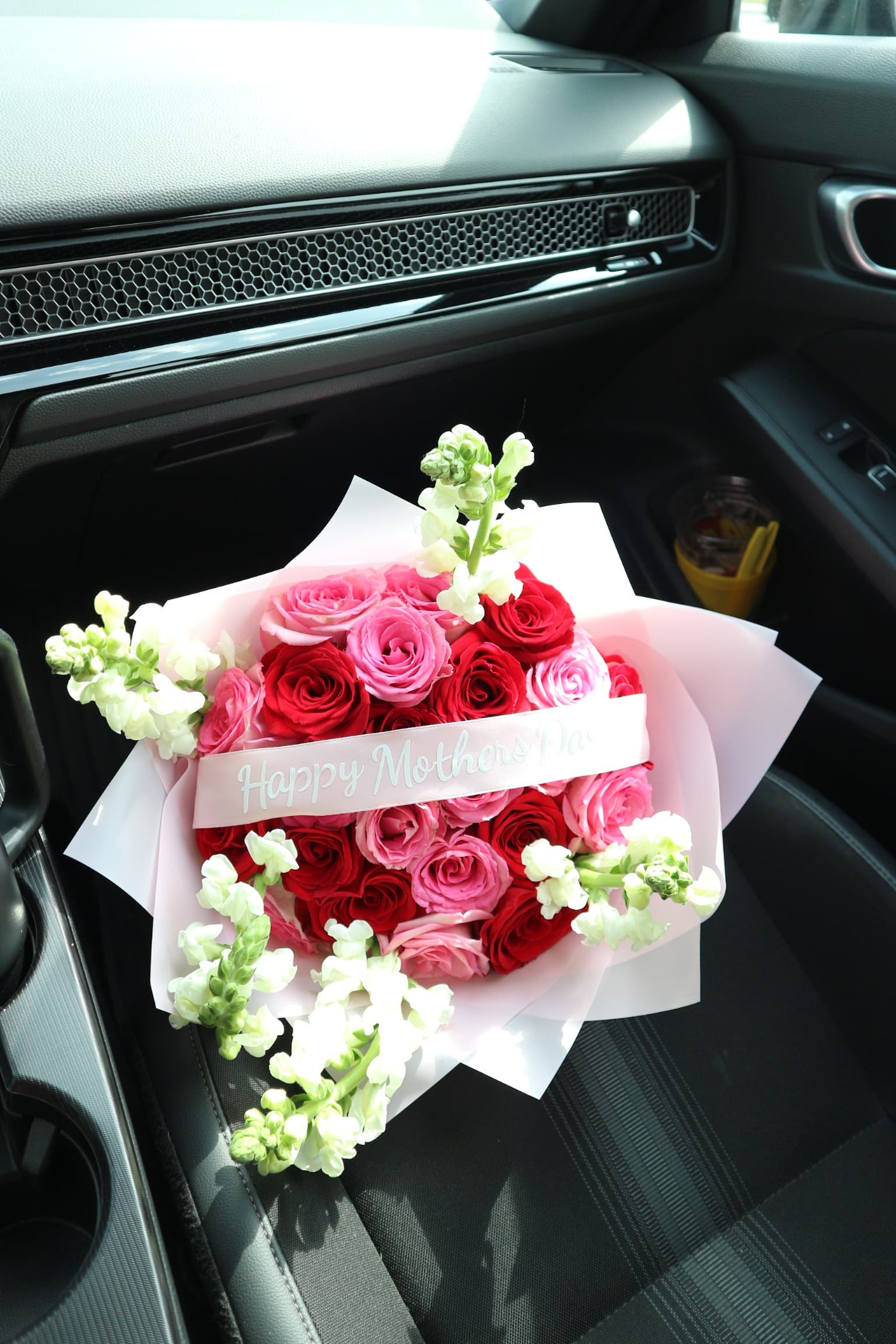 Red and pink roses with white stock in pink wrap, Happy Mother's Day ribbon, shown on a car seat