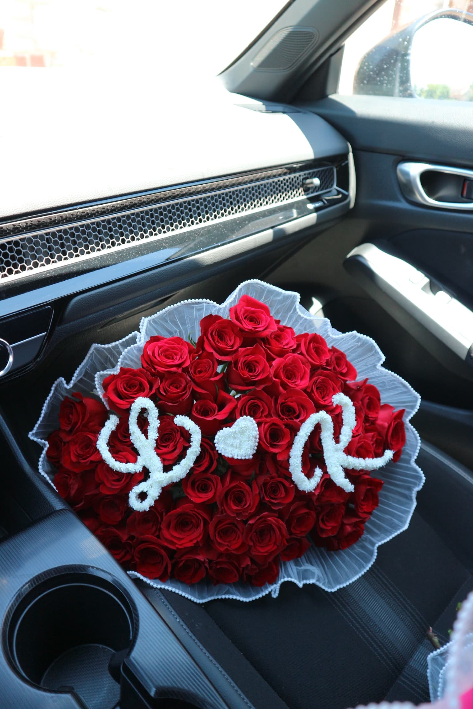 Red roses with pearl script initials and a heart, letter R visible, white ruffled wrap, in a car
