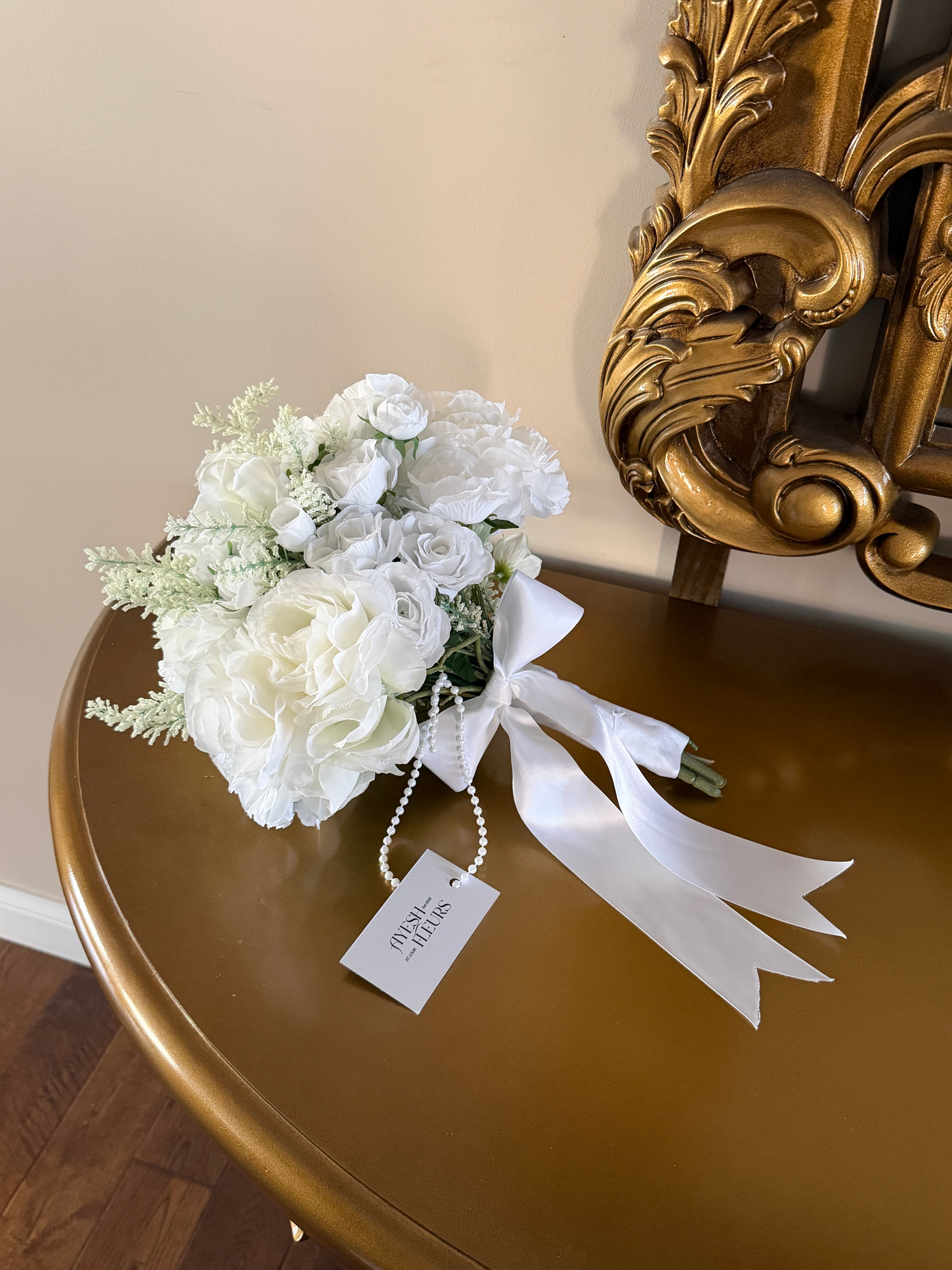 All-white mini bouquet of roses and peonies with satin bow and pearl strand on a gold table