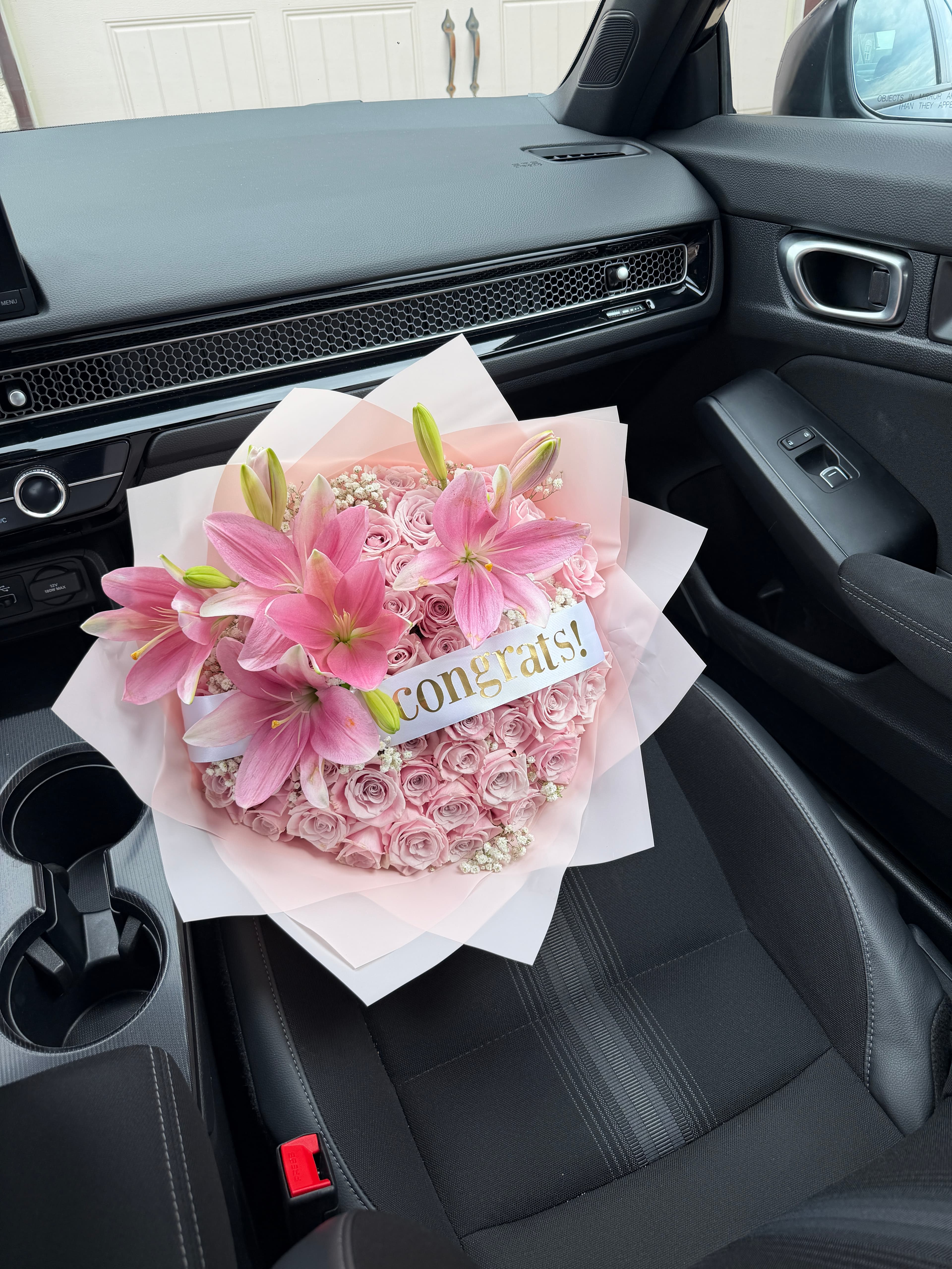 Pink lilies and blush roses with baby's breath in pink wrap and gold Congrats ribbon, in a car