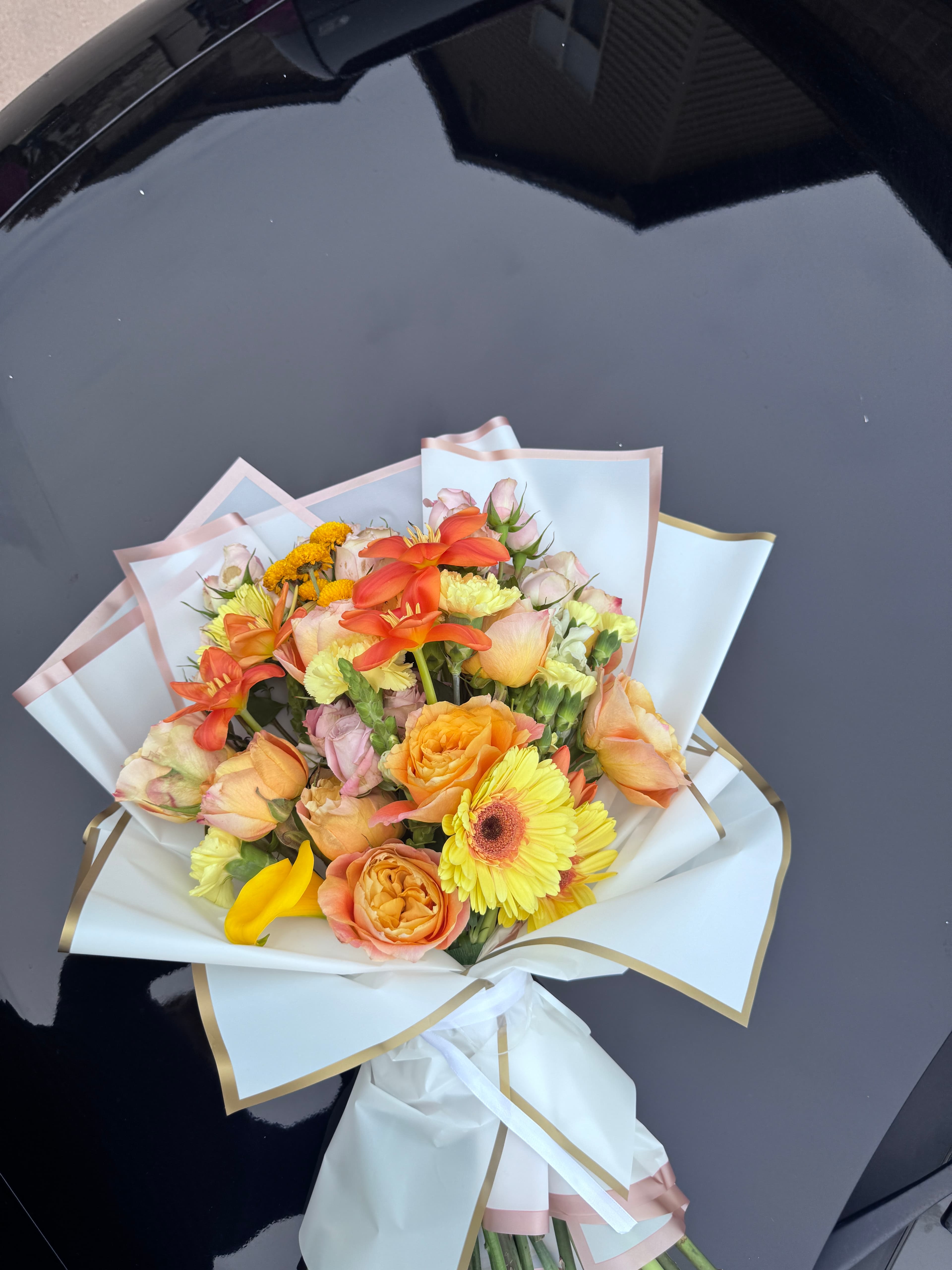 Orange and yellow bouquet of roses, gerbera daisies and coral lilies in white and gold wrap on a car hood