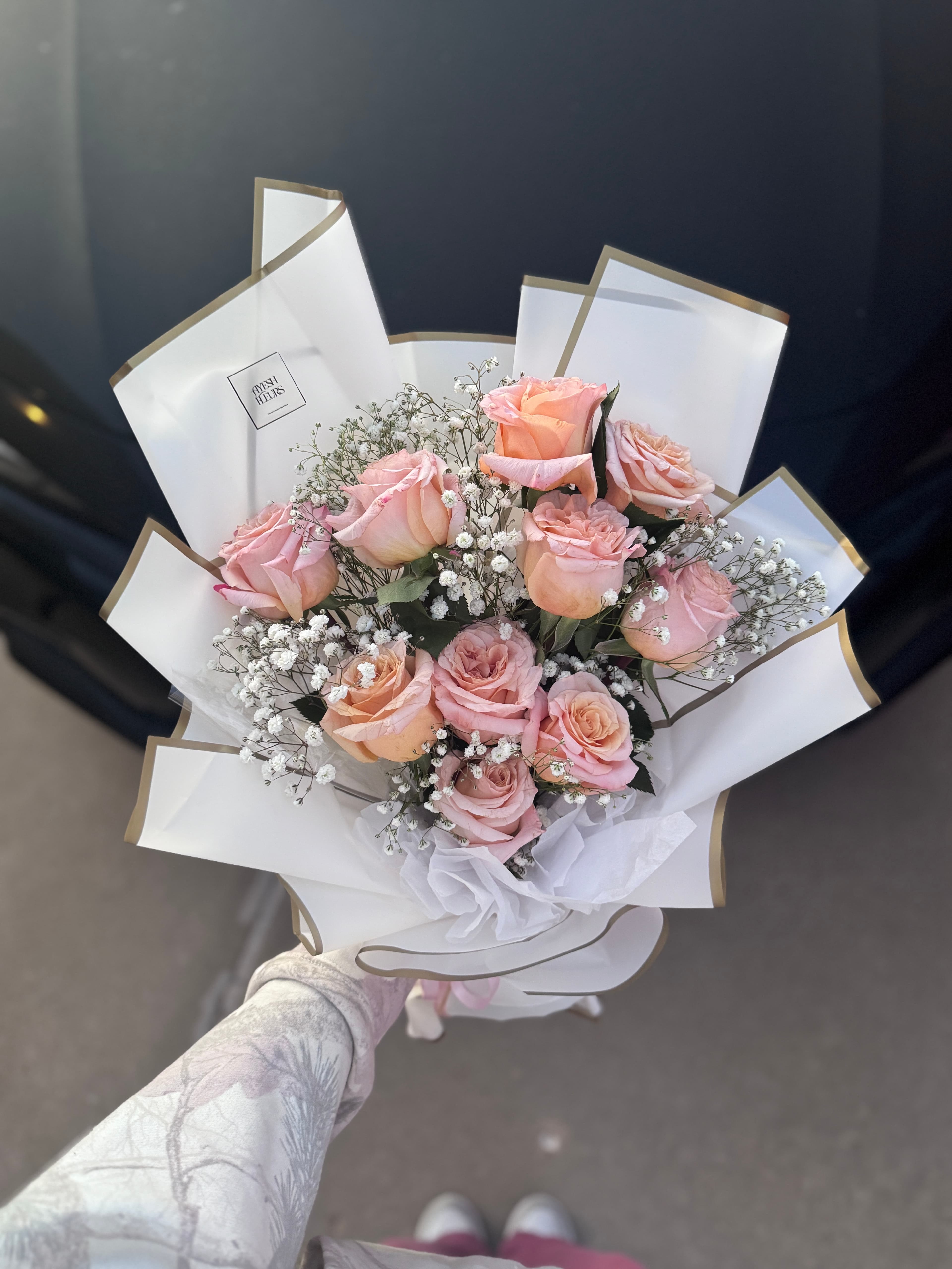 Blush pink roses with baby's breath in a white and gold petal wrap, held up outdoors