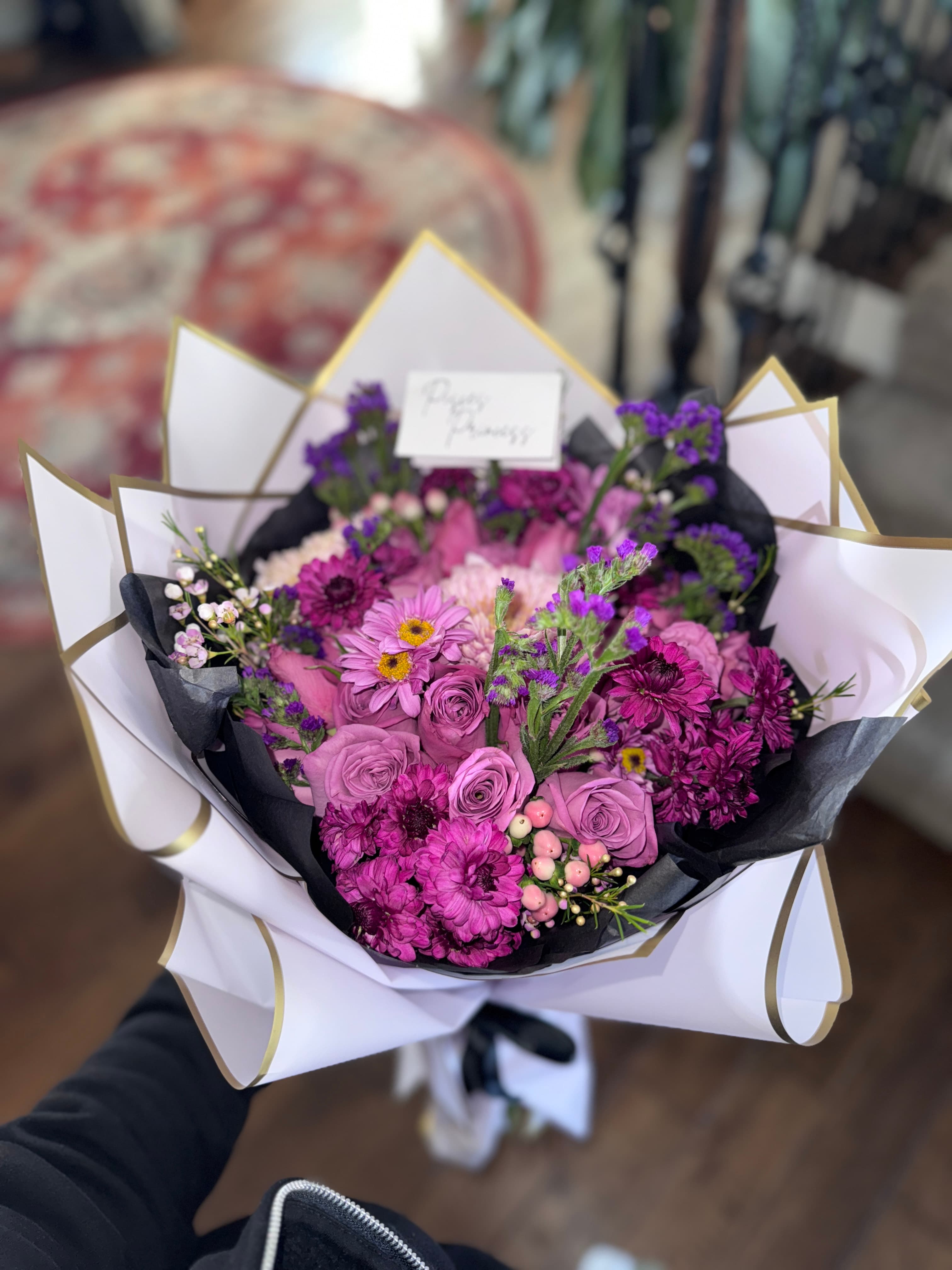 Purple bouquet of lavender roses, magenta daisies, statice and berries with a script card in black and white wrap