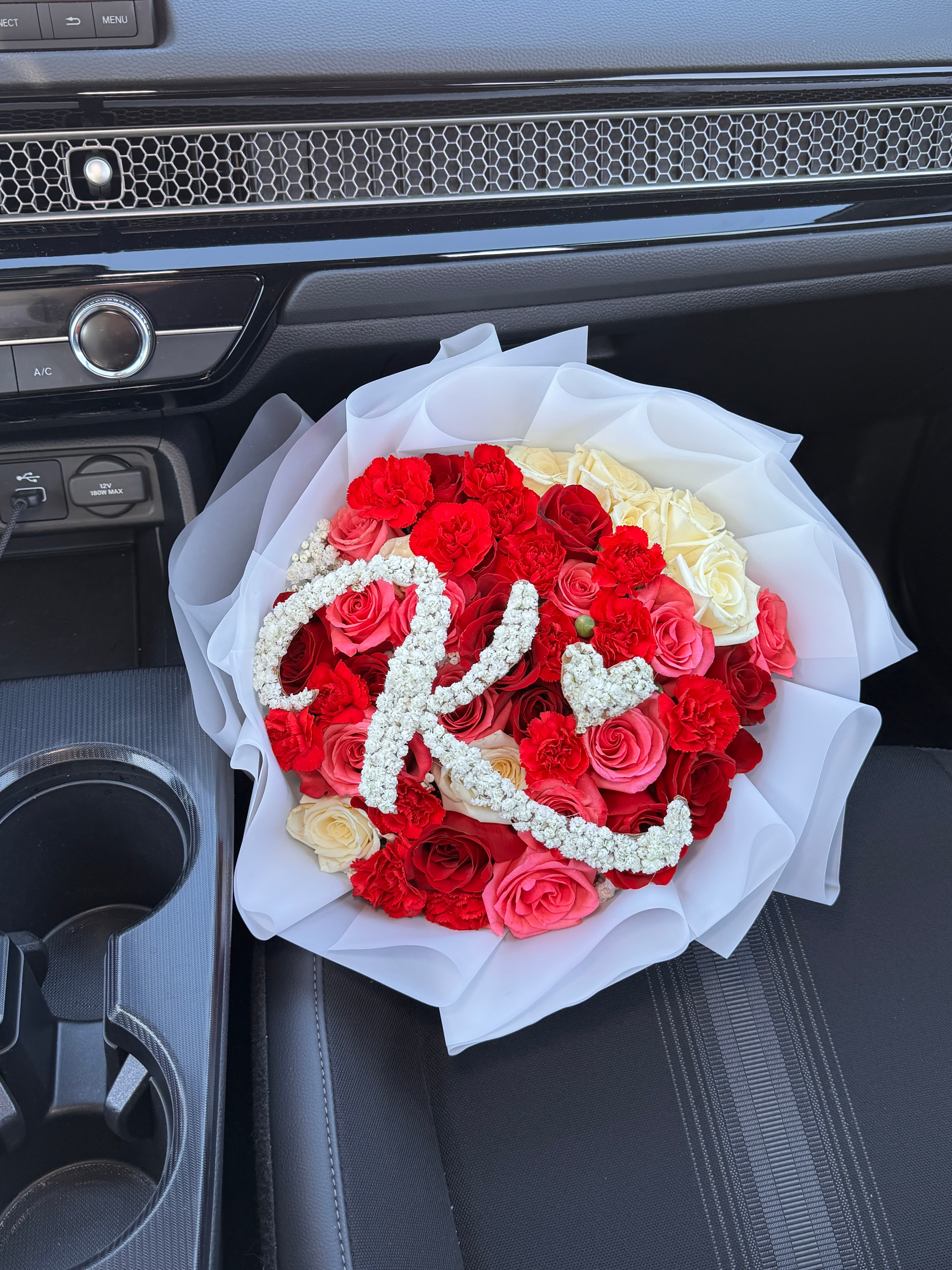 Red, pink and cream roses with an Arabic letter formed from tiny white blooms, white wrap, on a car seat