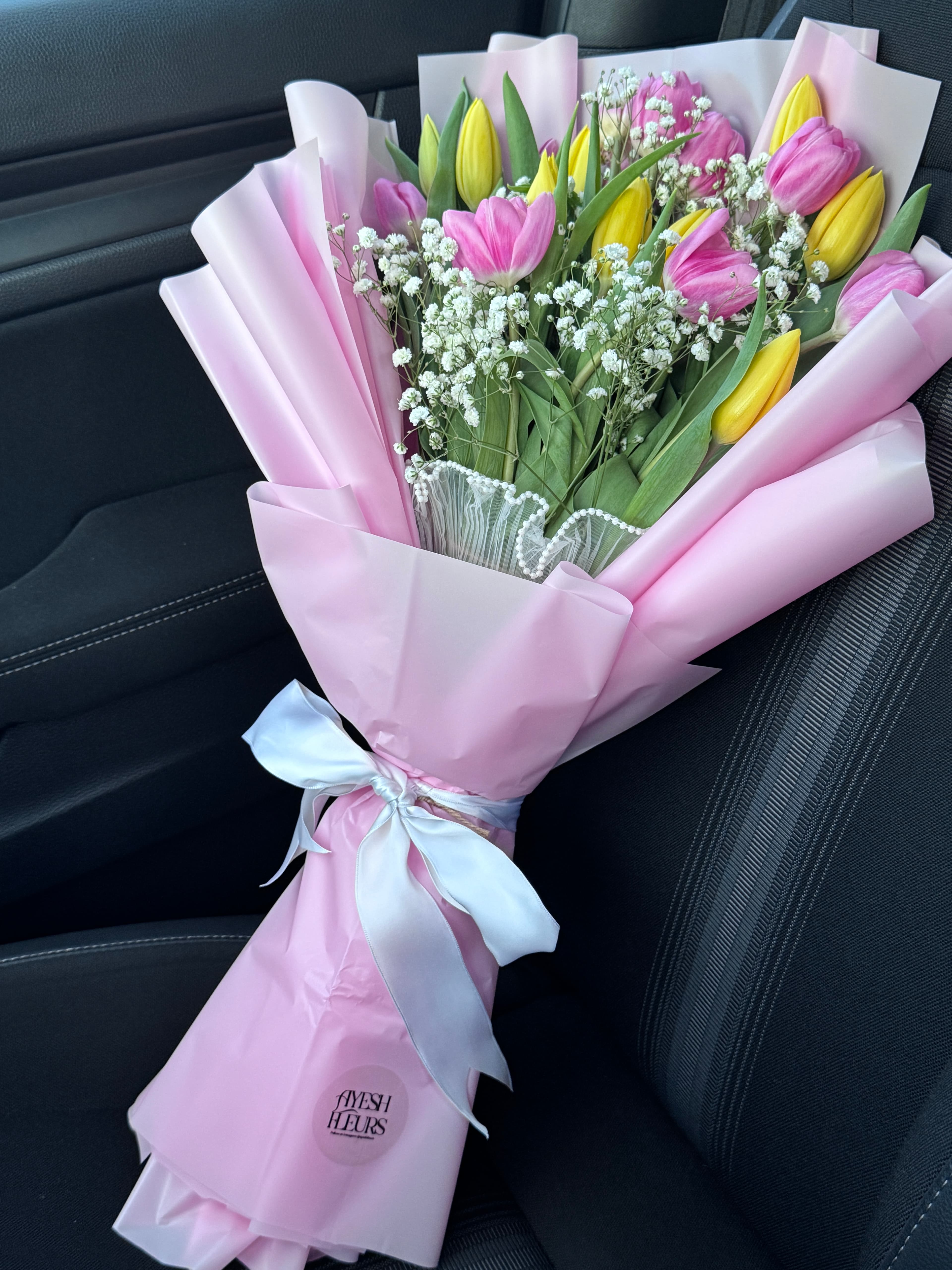 Pink and yellow tulips with baby's breath in pink wrap tied with a white ribbon bow, resting on a car seat