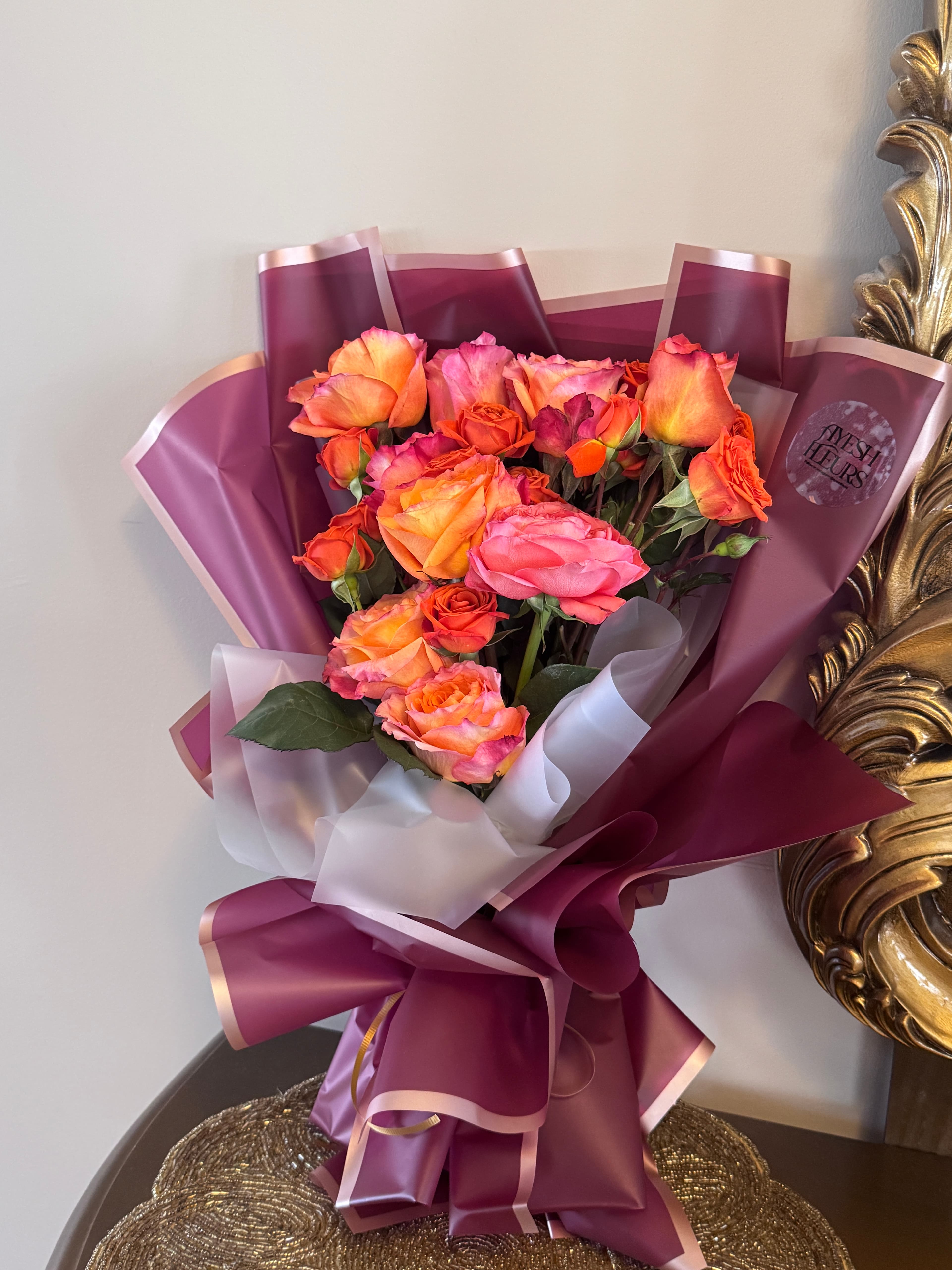 Orange and coral roses wrapped in burgundy and white paper, lying beneath an ornate gold mirror frame