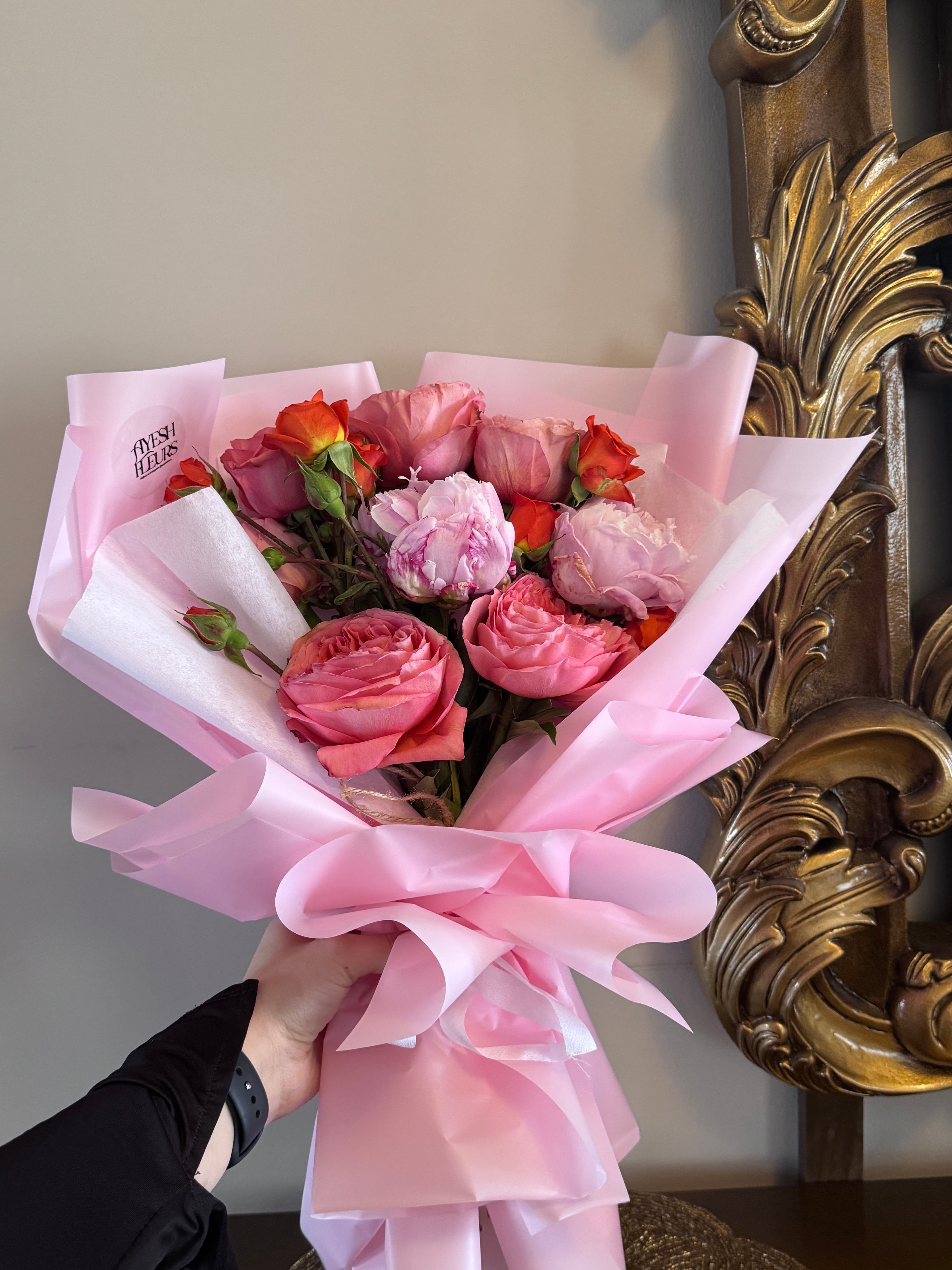 Coral garden roses, soft pink peonies and orange spray roses in pink wrap, held near a gold baroque mirror