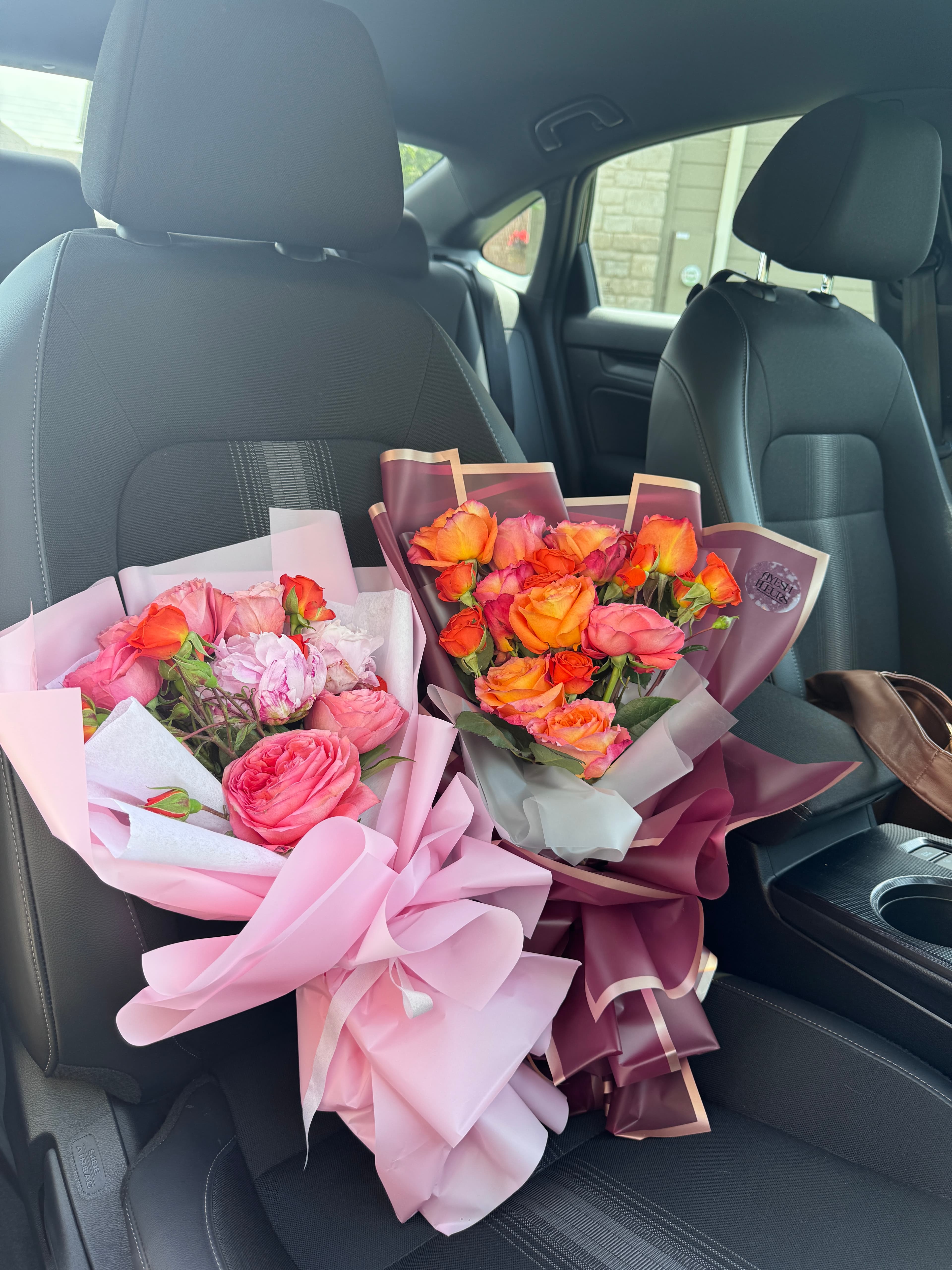 Two wrapped bouquets on a car seat: orange spray roses in burgundy wrap and pink roses in pale pink wrap