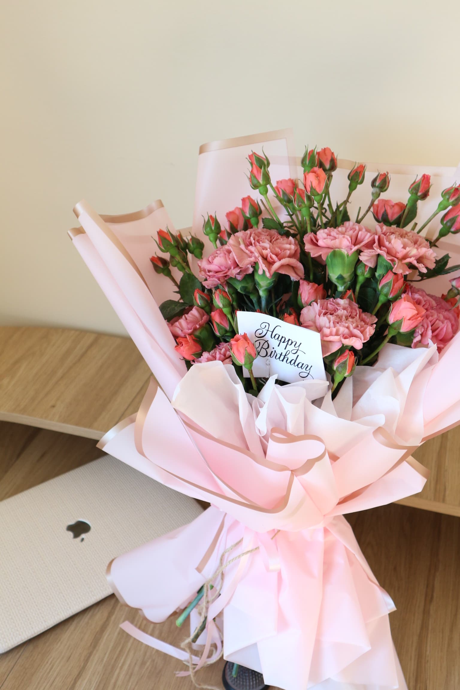 Pink carnations and coral spray roses in blush wrap with gold trim and a Happy Birthday card