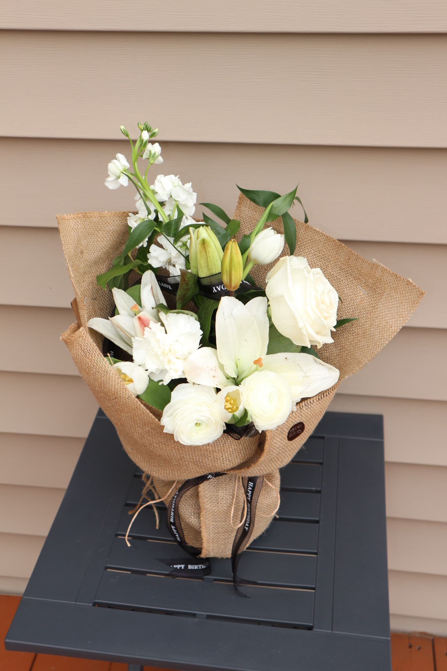 White lilies, roses, ranunculus and carnations in burlap wrap with a black Happy Birthday ribbon