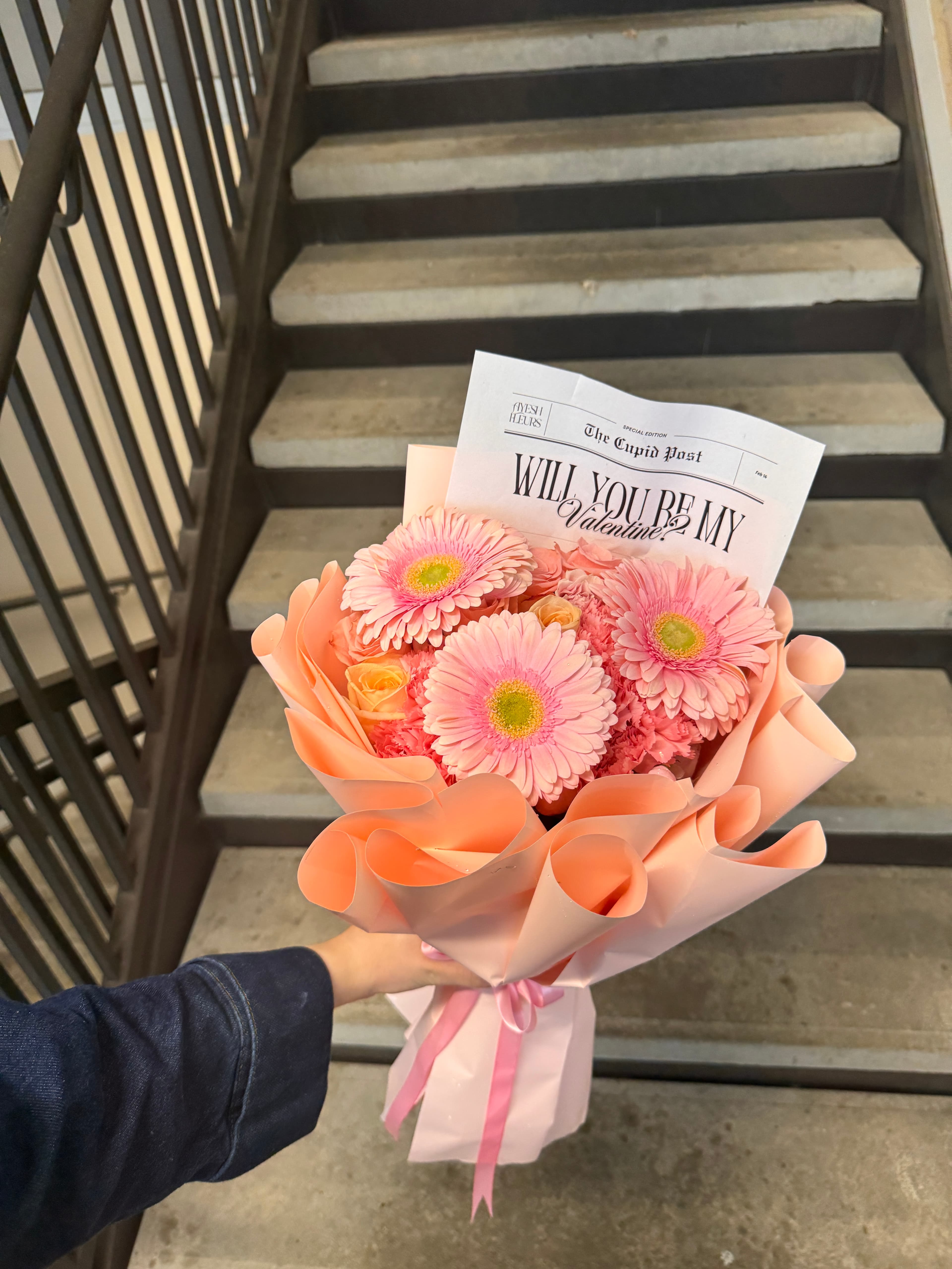 Pink gerbera daisies in peach ruffled wrap with a newspaper-style Will you be my card, held aloft