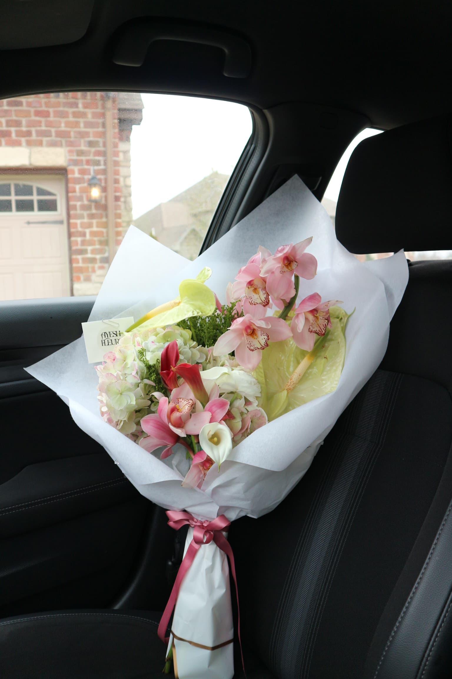Pink cymbidium orchids, calla lilies, hydrangea and green anthurium in white wrap on a car seat