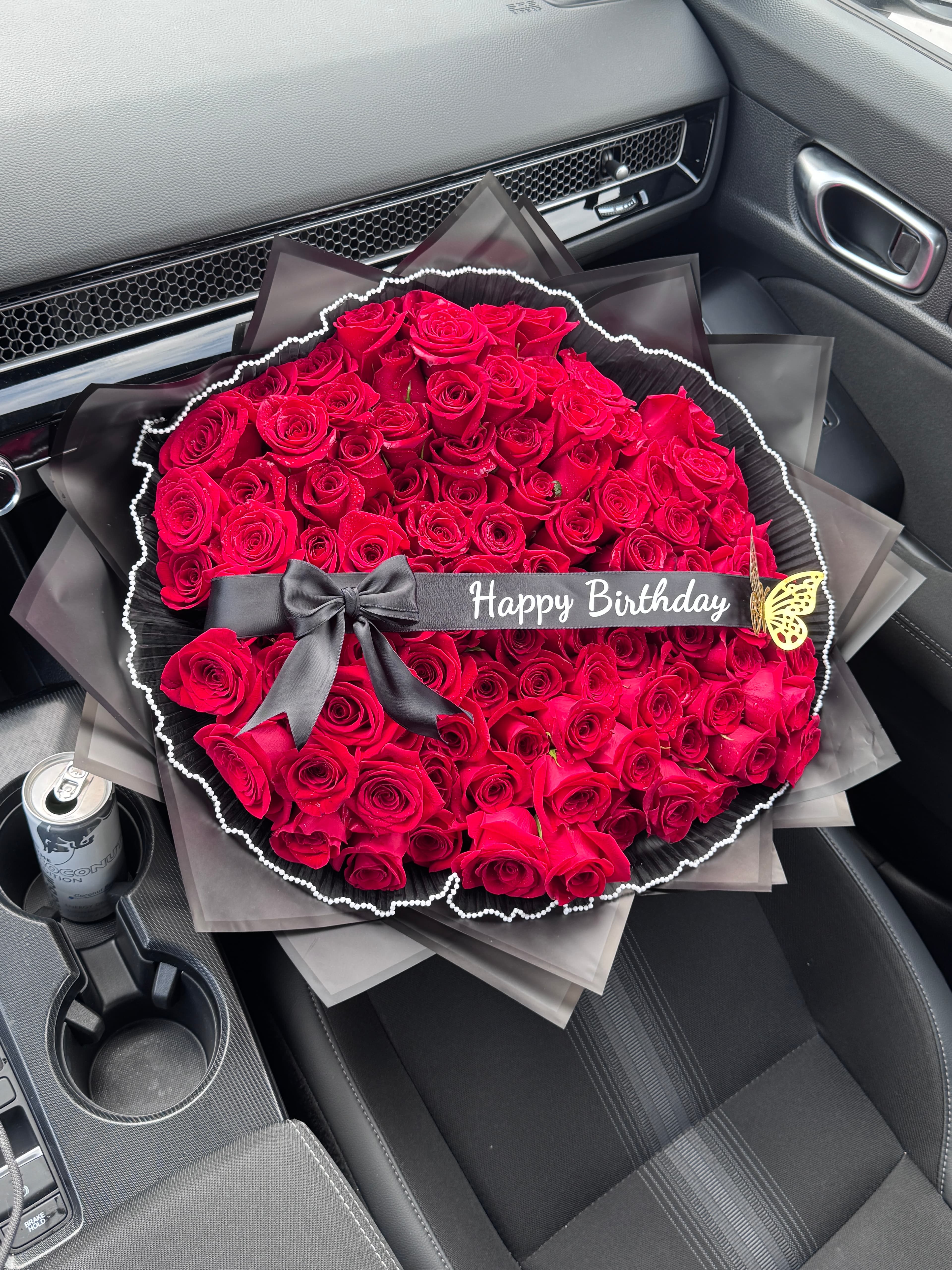 Red roses in black ruffled wrap with Happy Birthday ribbon, bow and gold butterfly on a car seat