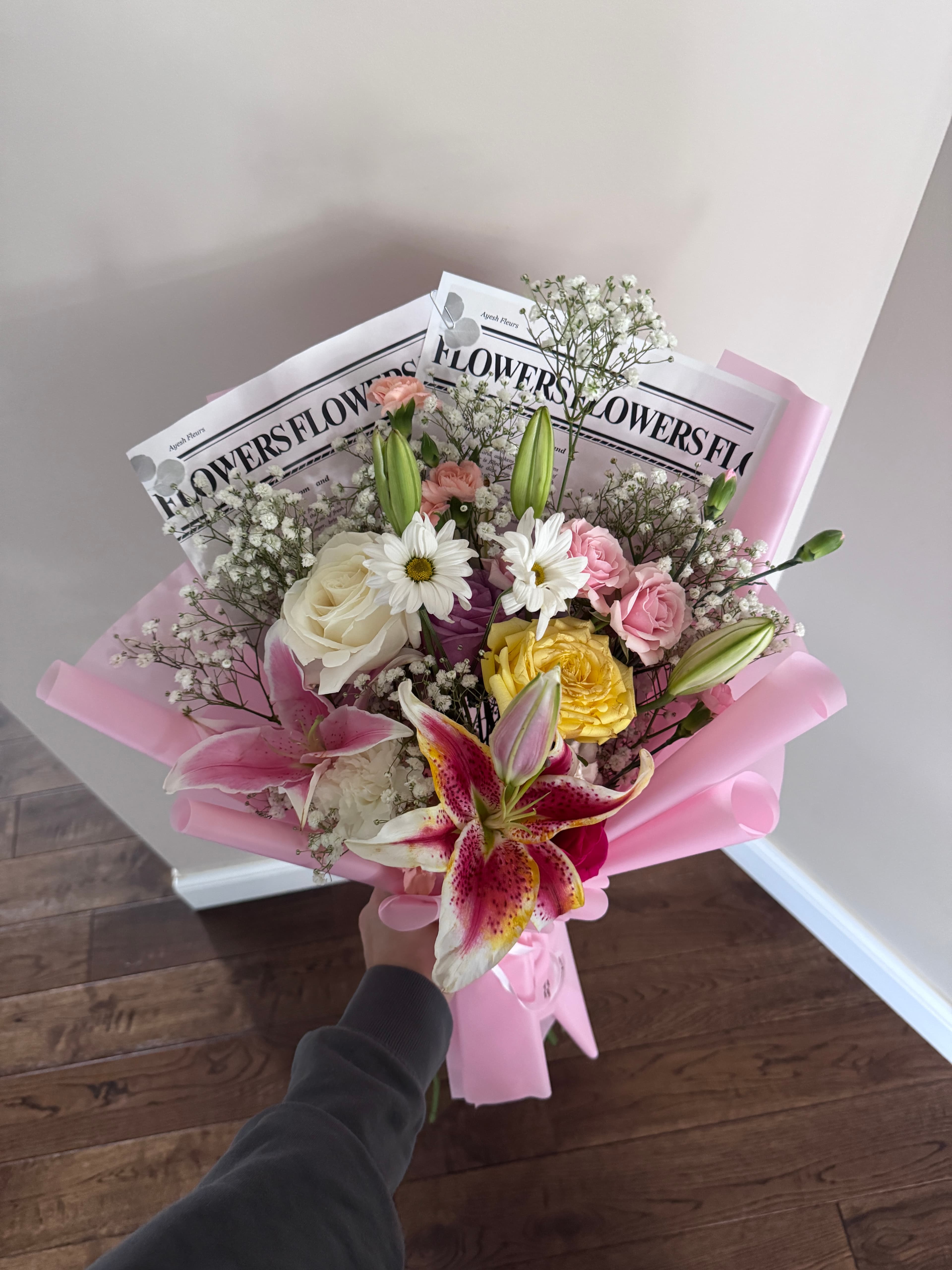 Stargazer lily, daisies and roses with baby's breath in pink wrap with Flowers newsprint, held by hand
