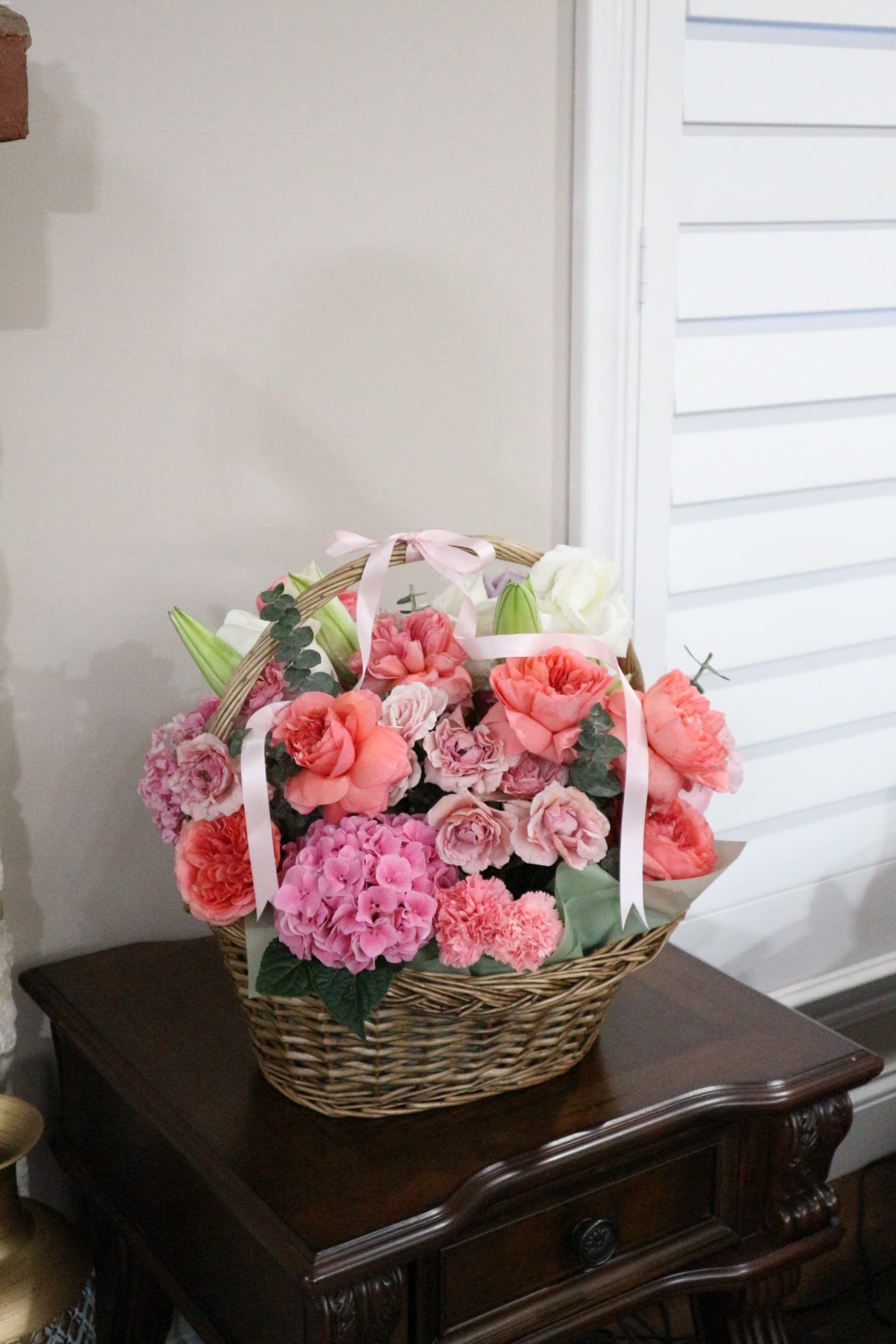Basket arrangement of pink hydrangea, coral garden roses, carnations and white lilies with pink ribbon