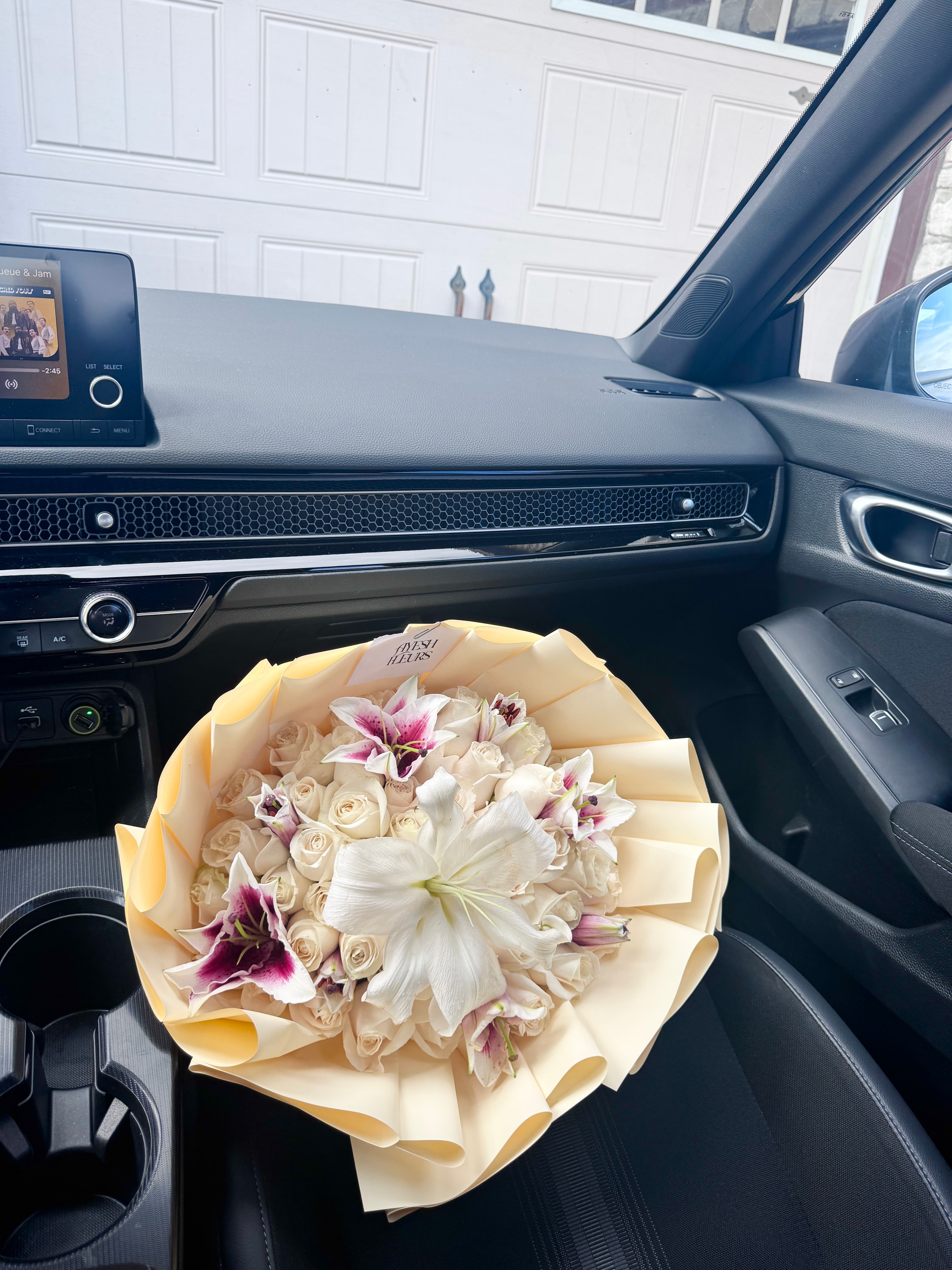 White roses and stargazer lilies in a ruffled cream wrap, resting inside a car