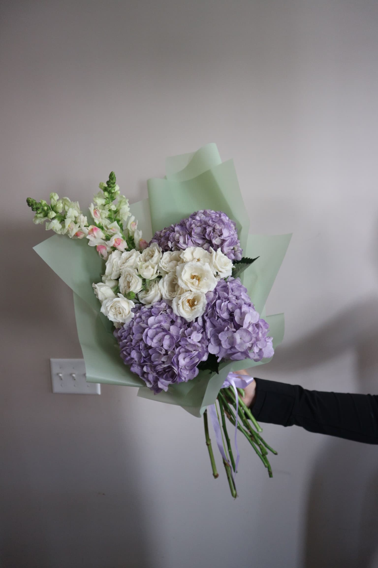 Purple hydrangea, white spray roses and snapdragons in sage green wrap with lilac ribbon, held aloft