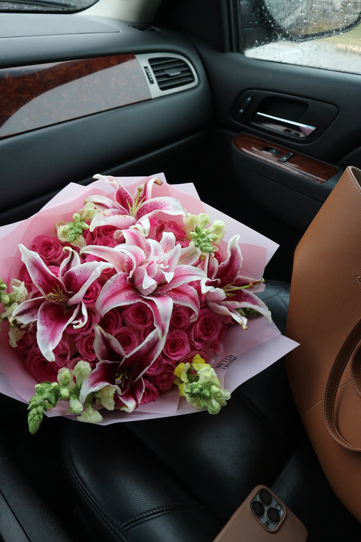 Hot pink roses packed with stargazer lilies and green snapdragons in soft pink wrap, seen inside a car